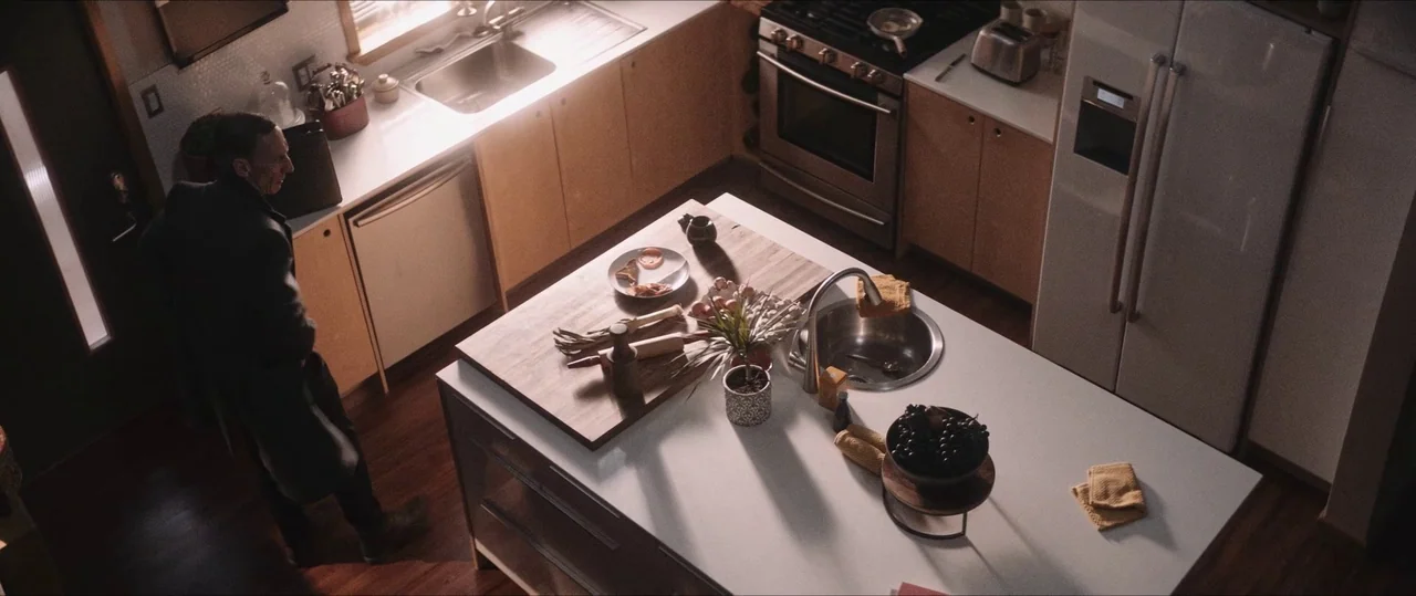 Anything For Jackson (2020) movie still: wide — A bright modern space with wooden cabinets and a central white stone island. A man stands silently…