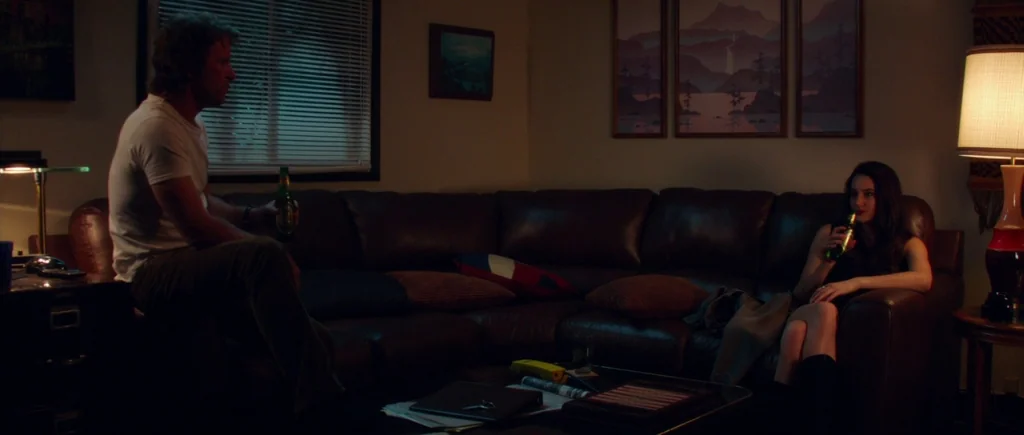 White Bird in a Blizzard (2014) movie still: wide — A dark, dimly lit living room features a large leather couch against a wall. A man sits on the…
