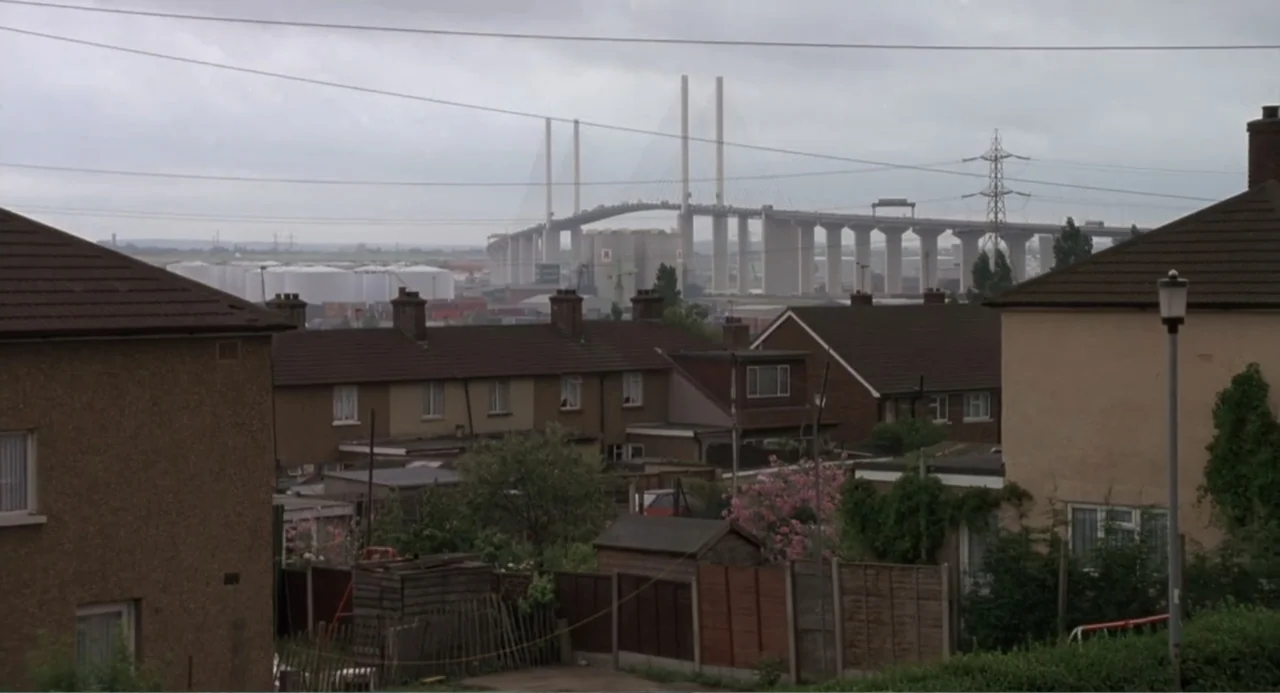Four Weddings and a Funeral (1994) movie still: establishing — Rows of suburban terraced houses occupy the foreground under a heavy overcast sky. In…