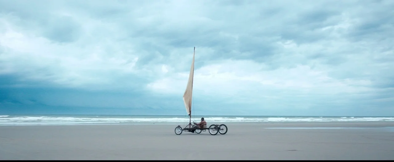 Slack Bay (2016) movie still: wide — A vast expanse of flat sand under a thick, grey, overcast sky. A person sits in a small, three-wheeled land…
