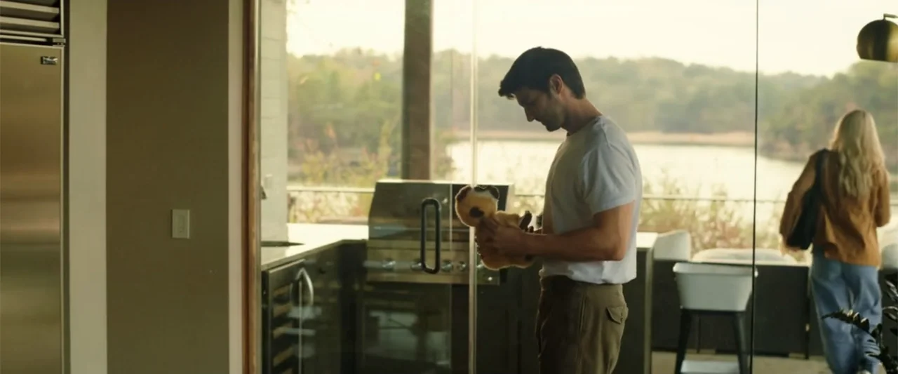 Someone Like You (2024) movie still: medium — A man stands inside a bright living space looking out toward a deck. He holds a small brown stuffed…