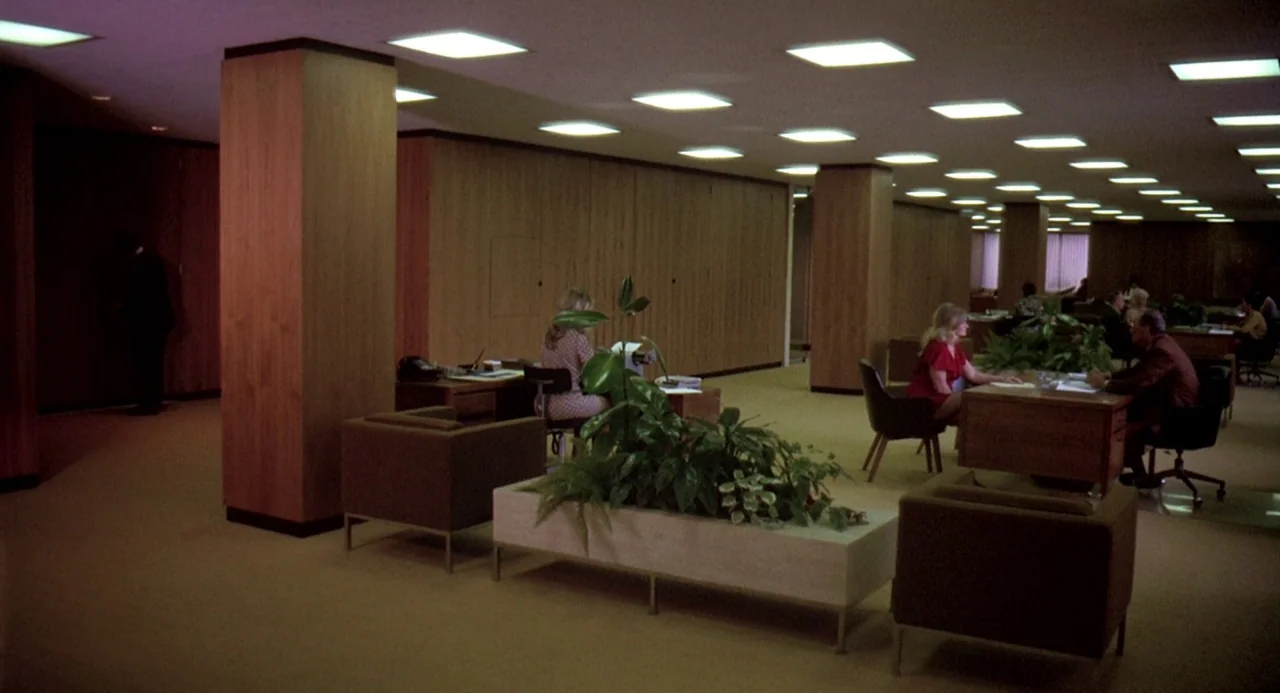 Charley Varrick (1973) movie still: wide — A spacious 1970s open-plan office features wood-paneled walls and rows of recessed ceiling lights.…