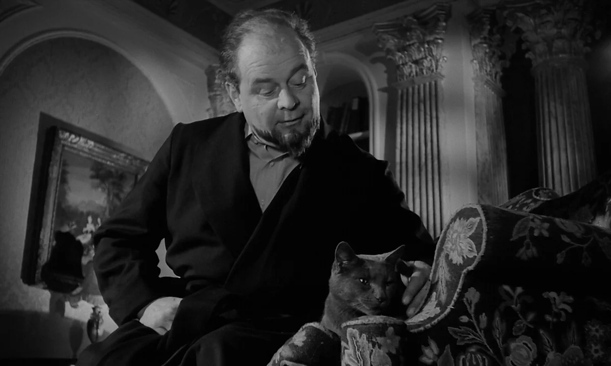 Night Of The Demon (1957) movie still: medium — A man sits in an armchair inside a room with classic columns and decor. He pets a cat while looking…
