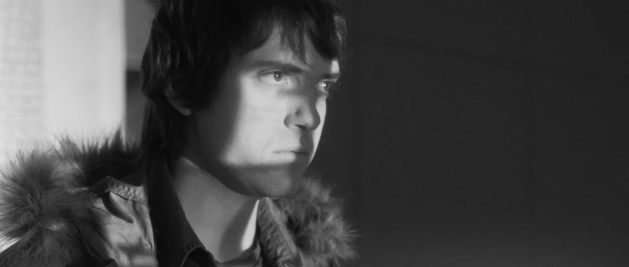 Polytechnique (2009) movie still: closeup — An interior space is dimly lit by a sharp side light source. A young man with dark shaggy hair stands…