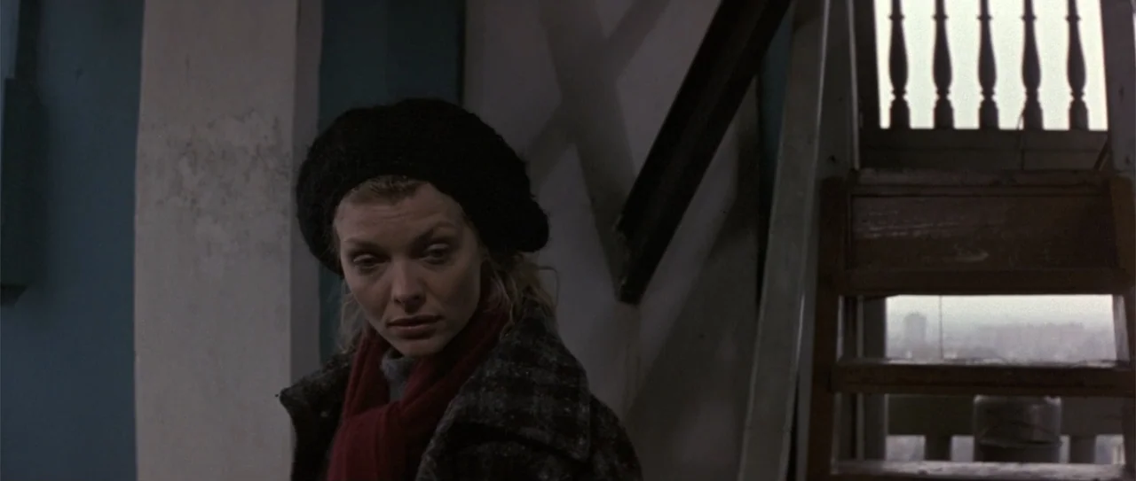 The Russia House (1990) movie still: medium — A dimly lit indoor stairwell features wooden stairs and structural columns. A woman in a dark coat and…