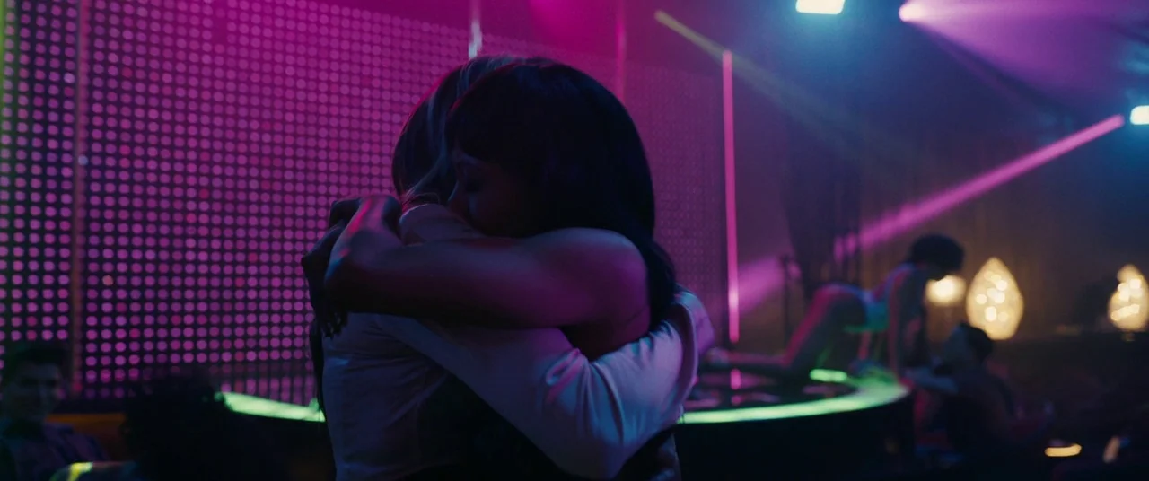 Hustlers (2019) movie still: medium — A dimly lit club interior features a circular stage with neon lighting and figures. Two people stand in the…
