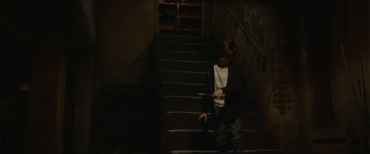 The Cellar (2022) movie still: fullbody — A dimly lit narrow stone staircase inside an old building with weathered walls and textures. A young boy…
