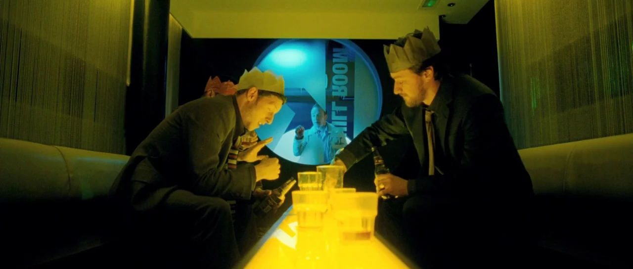 Filth (2013) movie still: medium — A dimly lit room with long leather benches and a glowing yellow table surface. Two men wearing paper crowns are…