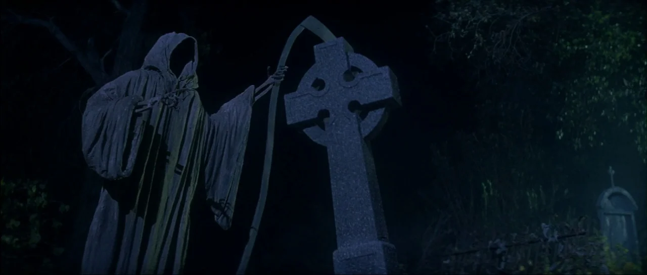 The Monster Squad (1987) movie still: medium — A dark, atmospheric graveyard at night features a stone cross tombstone near dense trees. A hooded,…