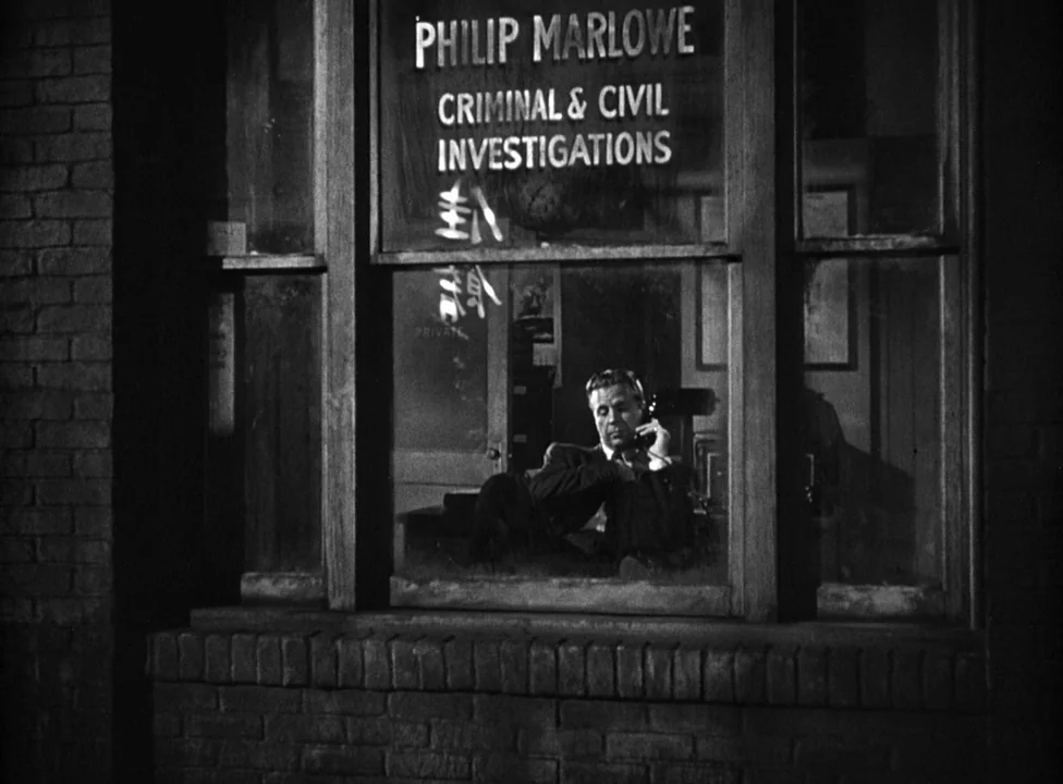 Murder, My Sweet (1944) movie still: medium — A brick exterior view looking through a window into a dark detective agency office. A man sits at his…