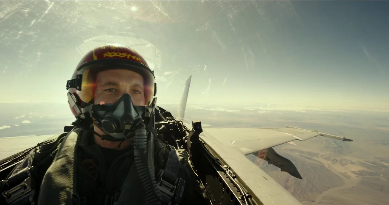 Top Gun: Maverick (2022) movie still: closeup — A fighter pilot sits in the cockpit of a jet flying over a desert. The aircraft wing is visible on…