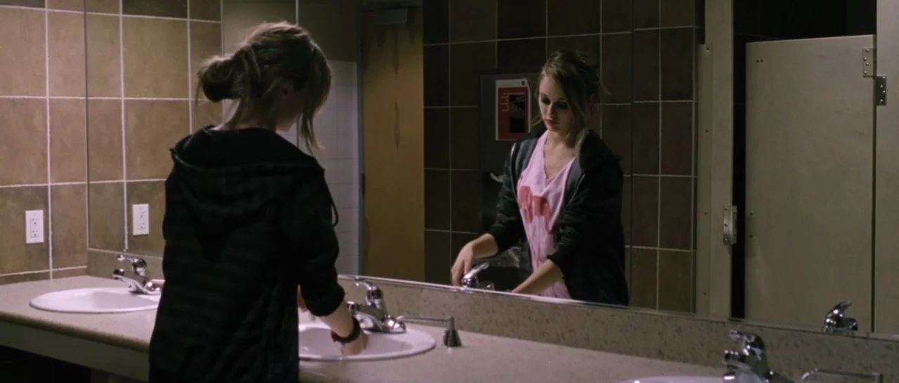 Wildlike (2014) movie still: medium — A public restroom features dark brown tiled walls and two ceramic sinks. A young woman stands at the counter…