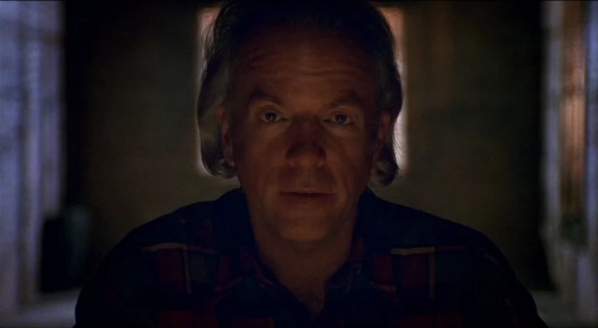 Gray’s Anatomy (1996) movie still: closeup — A man stands centered in a dimly lit, nondescript interior space. He is looking directly into the…