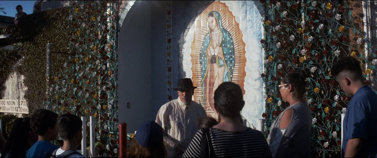El Chicano (2018) movie still: medium — An outdoor shrine features a large tile mosaic of the Virgin of Guadalupe. A group of people gathers as a…