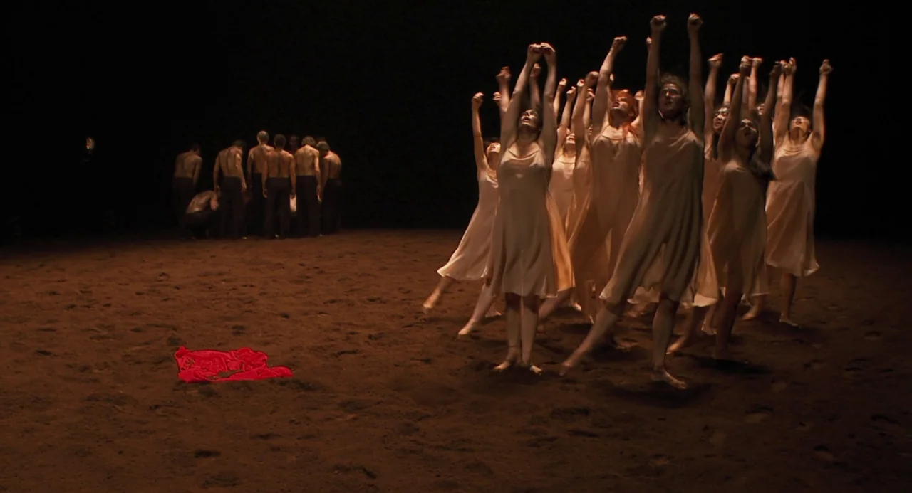 Pina (2011) movie still: wide — A dark space with a floor covered in dark brown loose earth or sand. A large group of dancers in pale dresses stands…