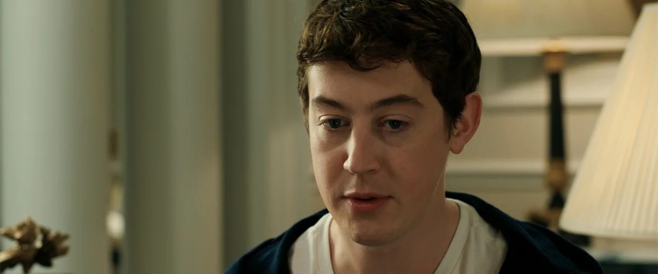 The Hustle (2019) movie still: closeup — A well-lit residential room features a lamp and neutral wall background. A young man with brown hair speaks…