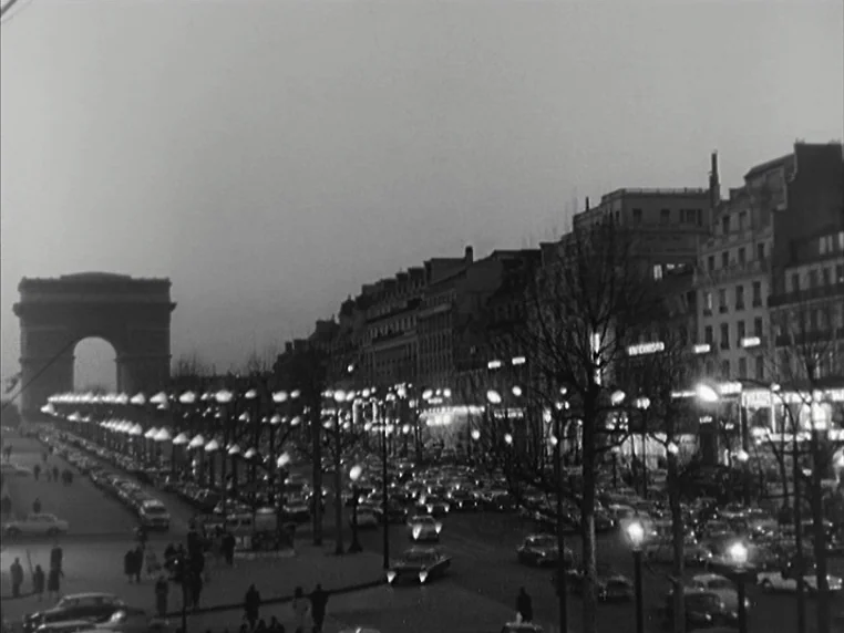 Vivre Sa Vie (1962) movie still: establishing — A wide view of a Parisian boulevard leads toward the Arc de Triomphe at night. Dense traffic fills…