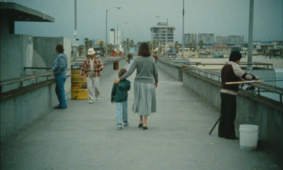 Documenteur (1981) movie still: fullbody — A concrete pier extends towards a sandy beach and city buildings under an overcast sky. A woman walks…