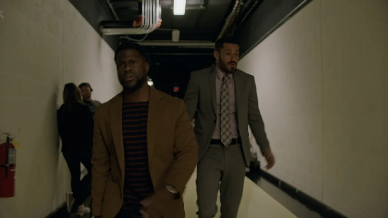 Die Hart (2023) movie still: medium — A narrow, institutional hallway with white walls and a dark ceiling. Two men walk side-by-side toward the…