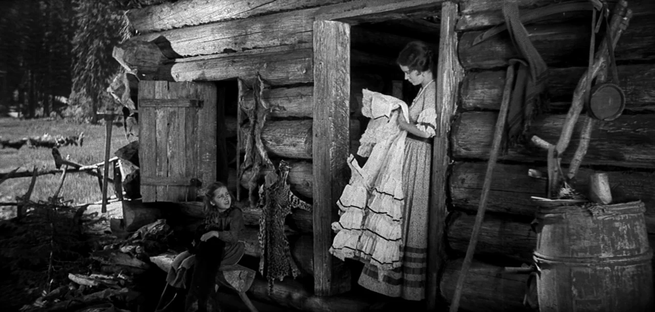 The Big Trail (1930) movie still: medium — A rustic log structure sits at the edge of a wooded area in daylight. A woman stands in the doorway…