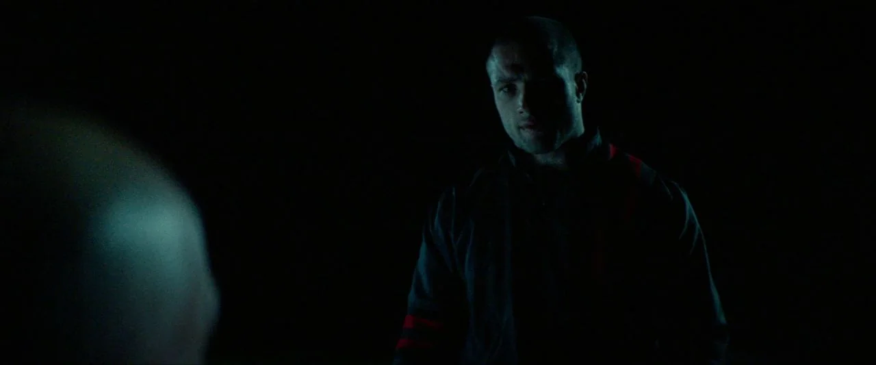 Calm With Horses (2019) movie still: medium — A dark, featureless environment surrounds a man in a striped athletic jacket. The man stands partially…