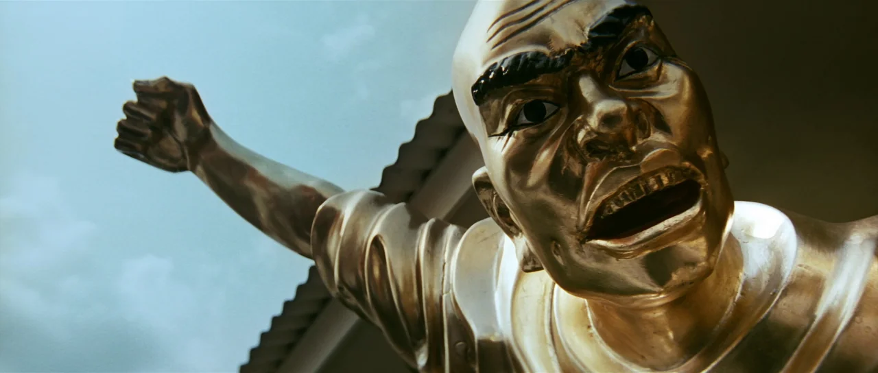 Infernal Affairs (2002) movie still: closeup — A shiny golden statue stands against a bright, clear blue sky background. The figure appears to be a…