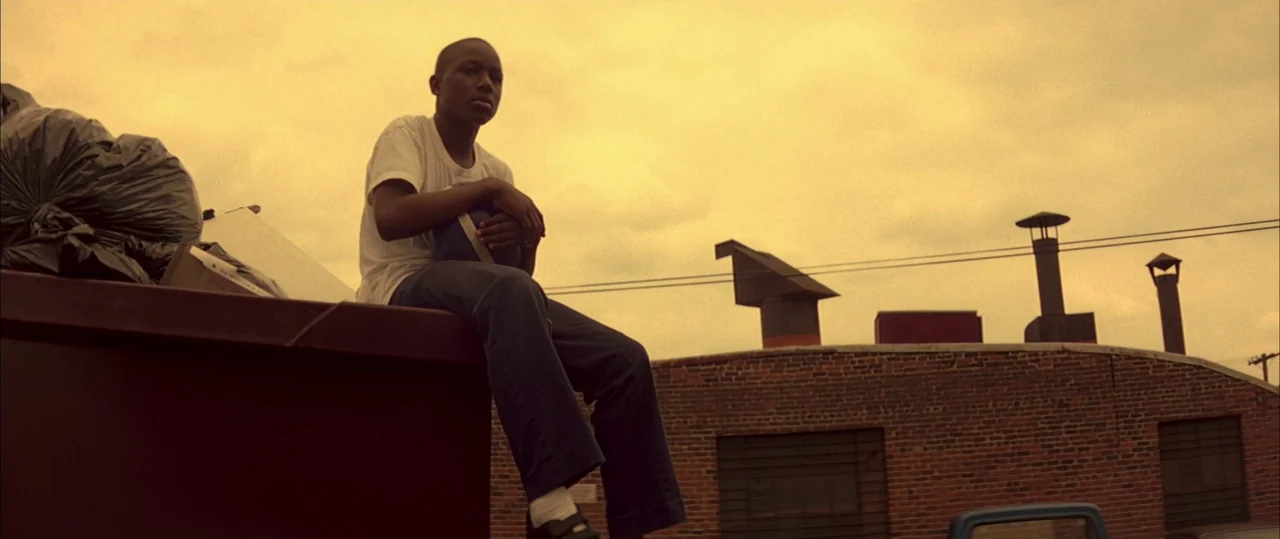 George Washington (2000) movie still: medium — A teenager sits on the edge of a large brown metal container. Behind him is a red brick building with…