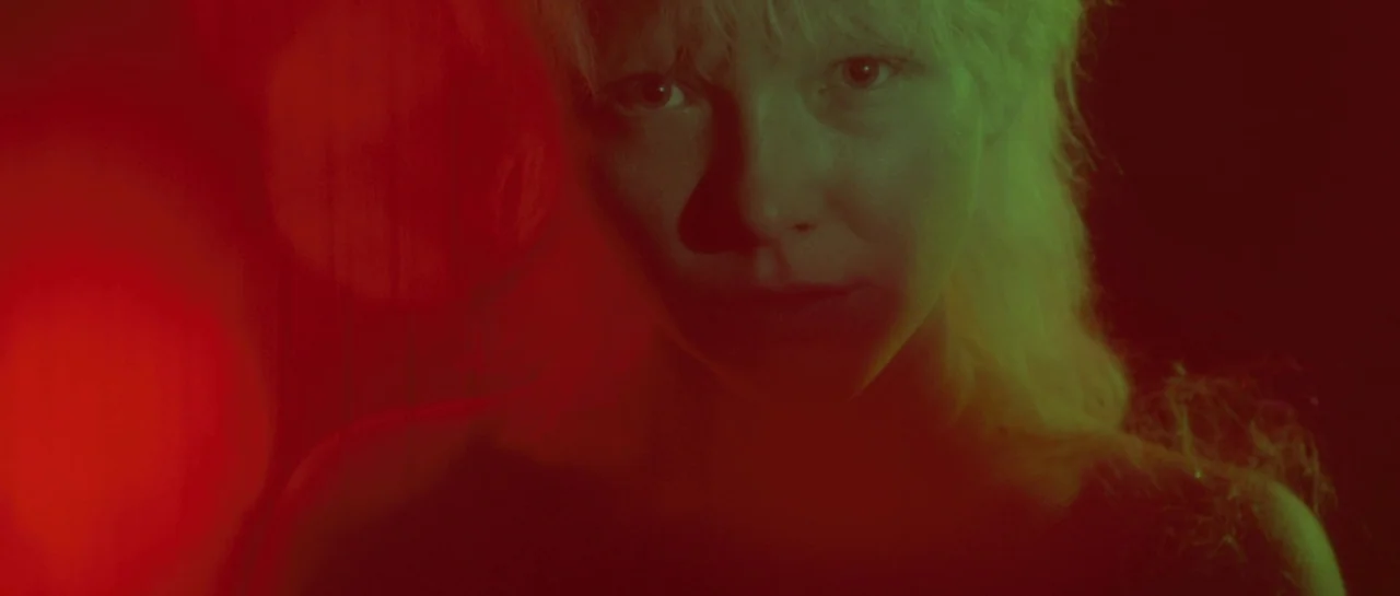 After Blue (2021) movie still: closeup — A dark, underexposed environment illuminated by colored light sources from either side. The subject stares…
