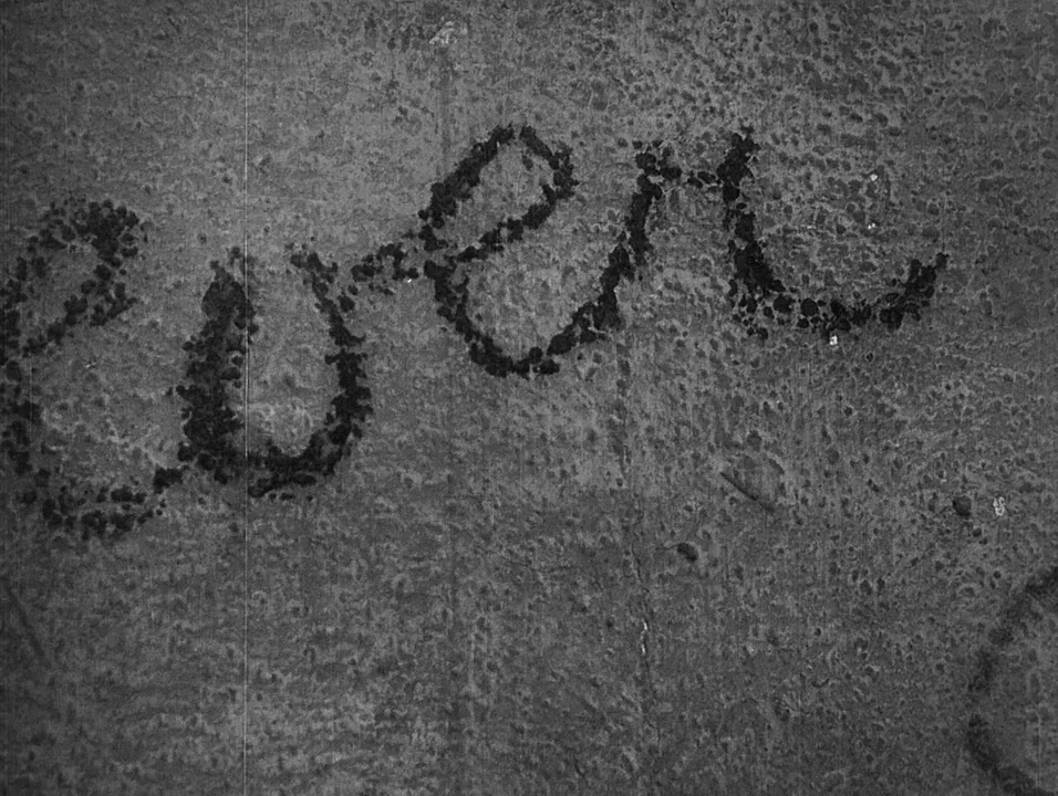Coeur Fidele (1923) movie still: closeup — A weathered, grainy concrete surface serves as a backdrop for dark hand-written cursive script. The…
