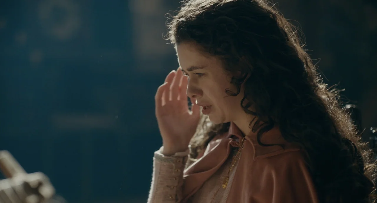Catherine Called Birdy (2022) movie still: medium — A dimly lit interior room with stone walls and soft directional light from a window. A young…