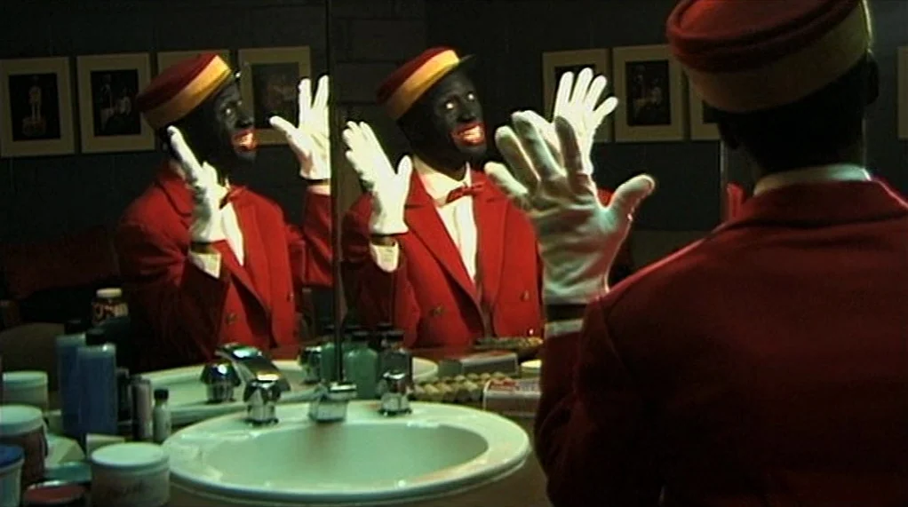 Bamboozled (2000) movie still: medium — A dark, dimly lit room contains a white ceramic sink and a wall mirror. The subject adjusts his hat and…