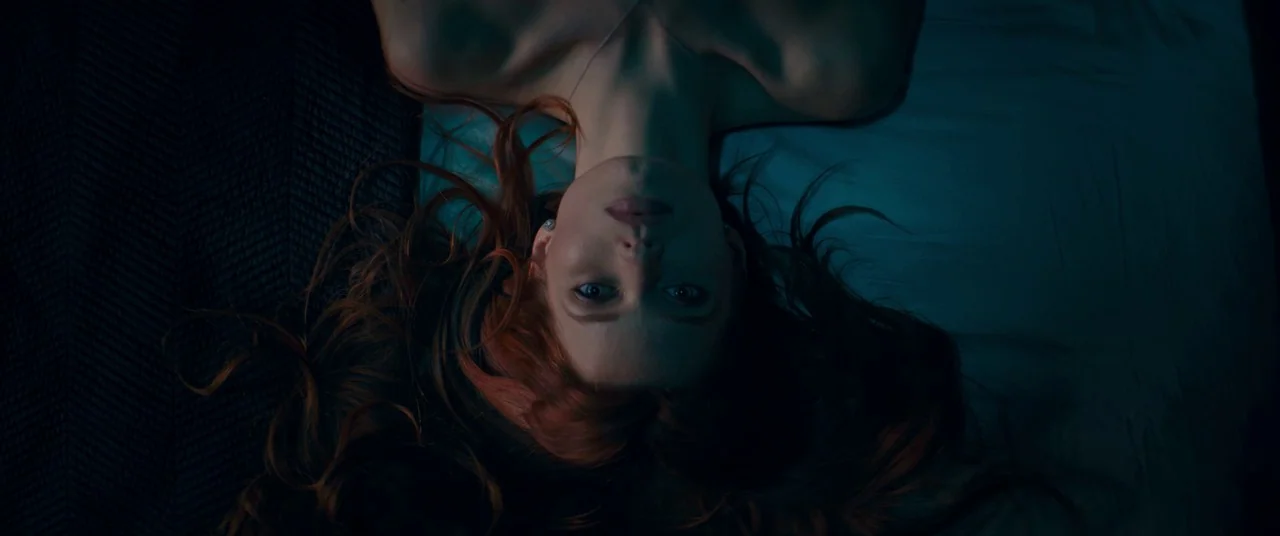 Elizabeth Harvest movie still: closeup — A woman lies on a surface covered in smooth, dark sheets. She faces directly toward the camera with her…