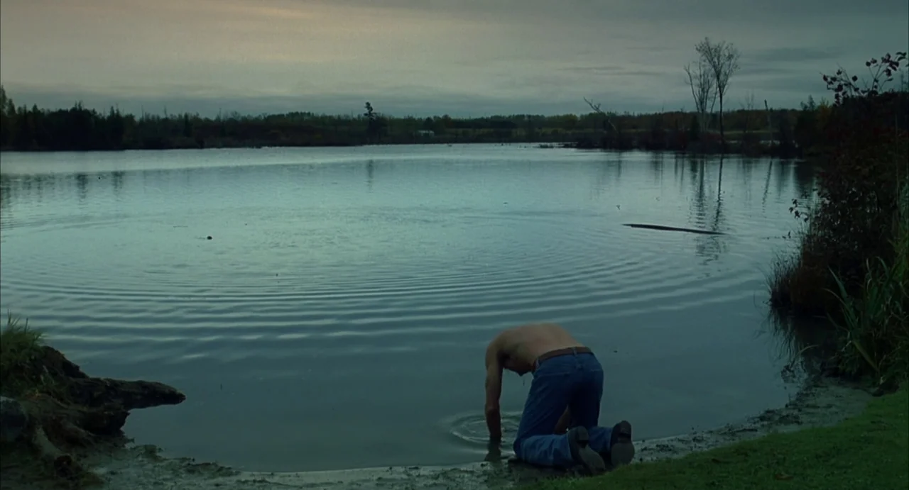 A History of Violence (2005) movie still: wide — A man kneels on the muddy bank of a still lake under an overcast sky. He reaches down with one hand…