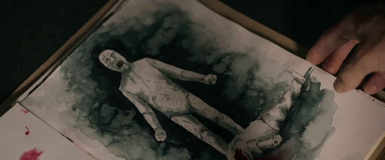 Oddity (2024) movie still: closeup — A hand holds an open sketchbook showing a dark watercolor painting of a figure. The painting displays a pale,…