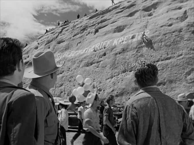 Ace In The Hole (1951) movie still: wide — A crowd of people gathers at the base of a massive stone cliff wall. Painted white letters on the rock…