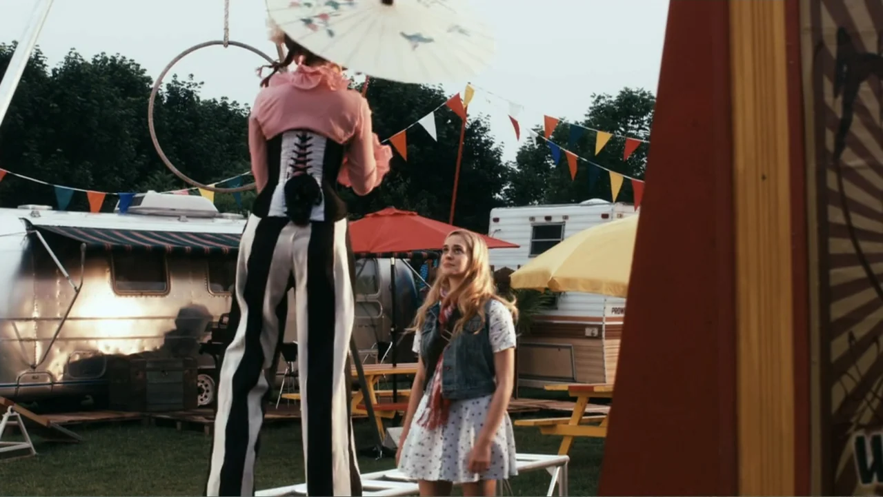 Sophie & Sheba (2010) movie still: medium — A carnival lot is filled with travel trailers and colorful bunting flags. A performer on tall stilts…
