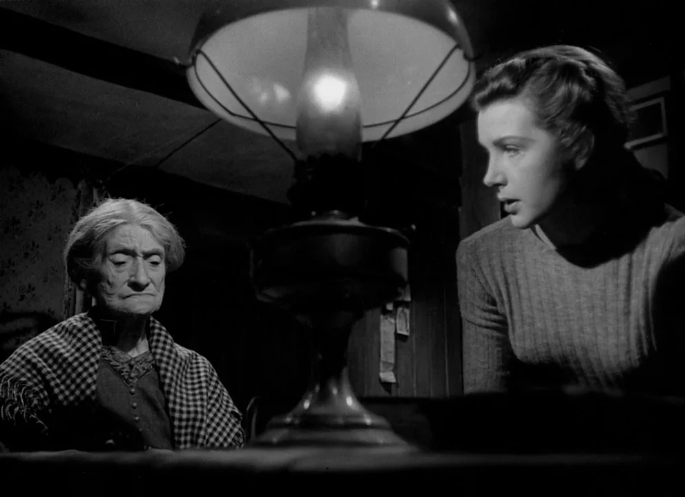 Odd Man Out (1947) movie still: medium — A dim room contains an old woman and a young woman separated by a lamp. The two women sit at a table facing…