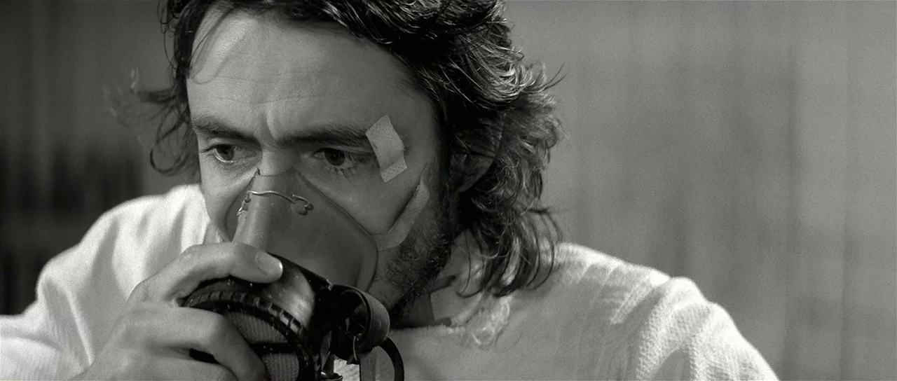 The Last Battle (1983) movie still: closeup — A man with long hair wears a medical mask in a dimly lit room. He holds the mask against his face with…