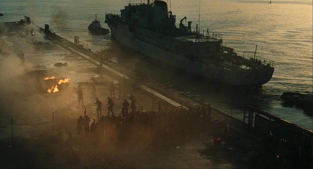 This Night (2008) movie still: establishing — A hazy, industrial harbor at sunset features a large naval vessel docked at a pier. A crowd of people…