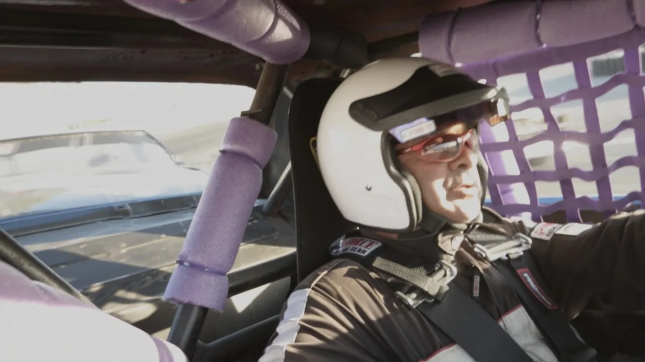 The Last Race (2022) movie still: medium — A racing car cockpit features purple foam padding on the roll cage and safety netting. A man wearing a…