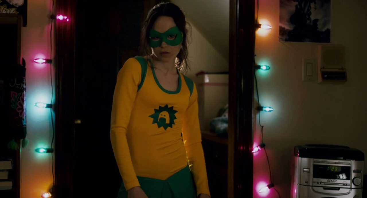 Super (2010) movie still: medium — A woman stands in a dimly lit doorway framed by decorative multi-colored holiday lights. She wears a superhero…
