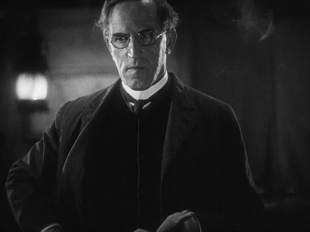 The Docks of New York (1928) movie still: medium — A dark, dimly lit room contains a man standing near a glowing lamp. The man wears formal 1920s…