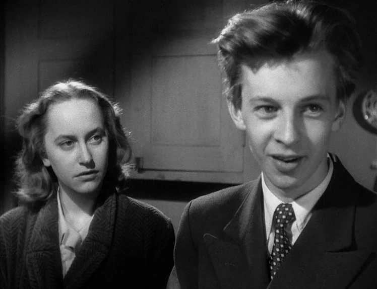 Prison (1949) movie still: medium — A dimly lit room with plain walls frames two individuals in close proximity. A young man talks while a woman…
