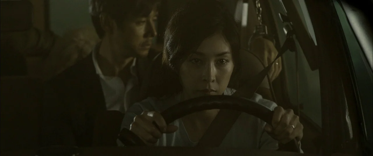 Creepy (2016) movie still: medium — The interior of a car at night with a woman driving. A man sits in the dark backseat with one hand raised…