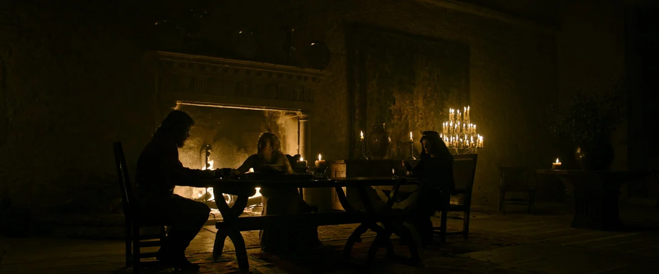 The Last Duel (2021) movie still: wide — A stone-walled medieval hall features a large burning fireplace and a long dining table. Three figures sit…