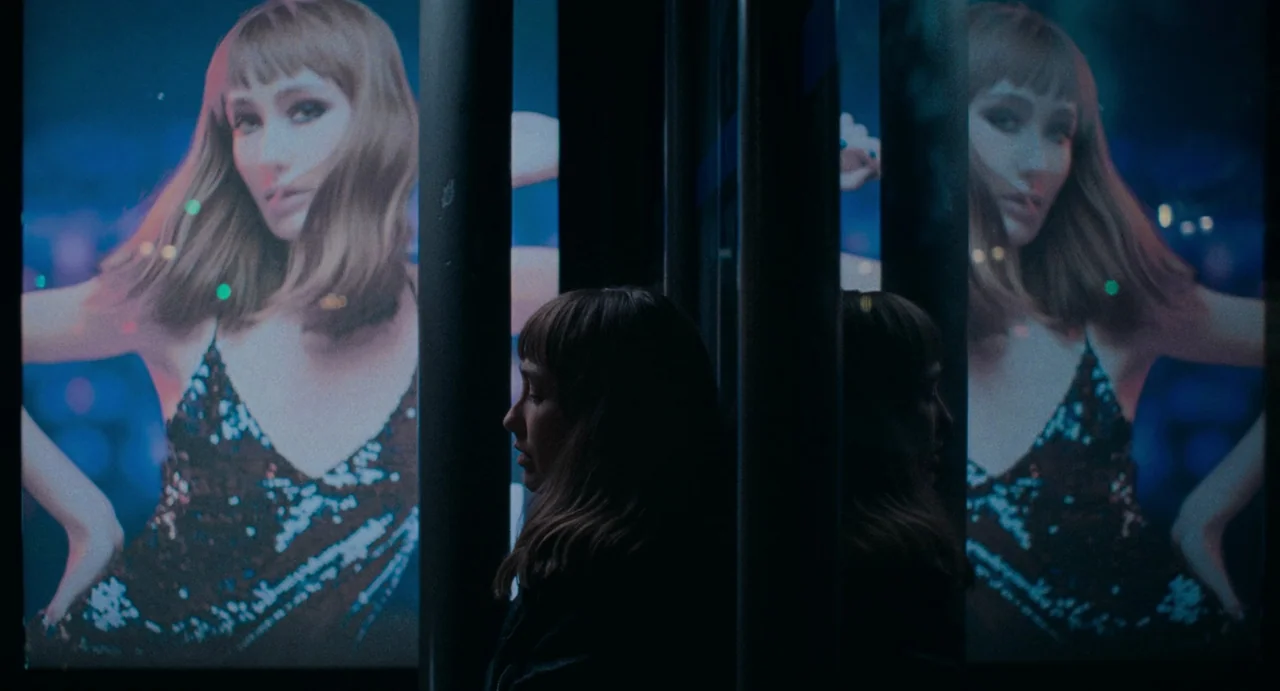 La Abuela (2021) movie still: medium — A woman stands between two illuminated advertising displays featuring a portrait of a woman. Dark vertical…