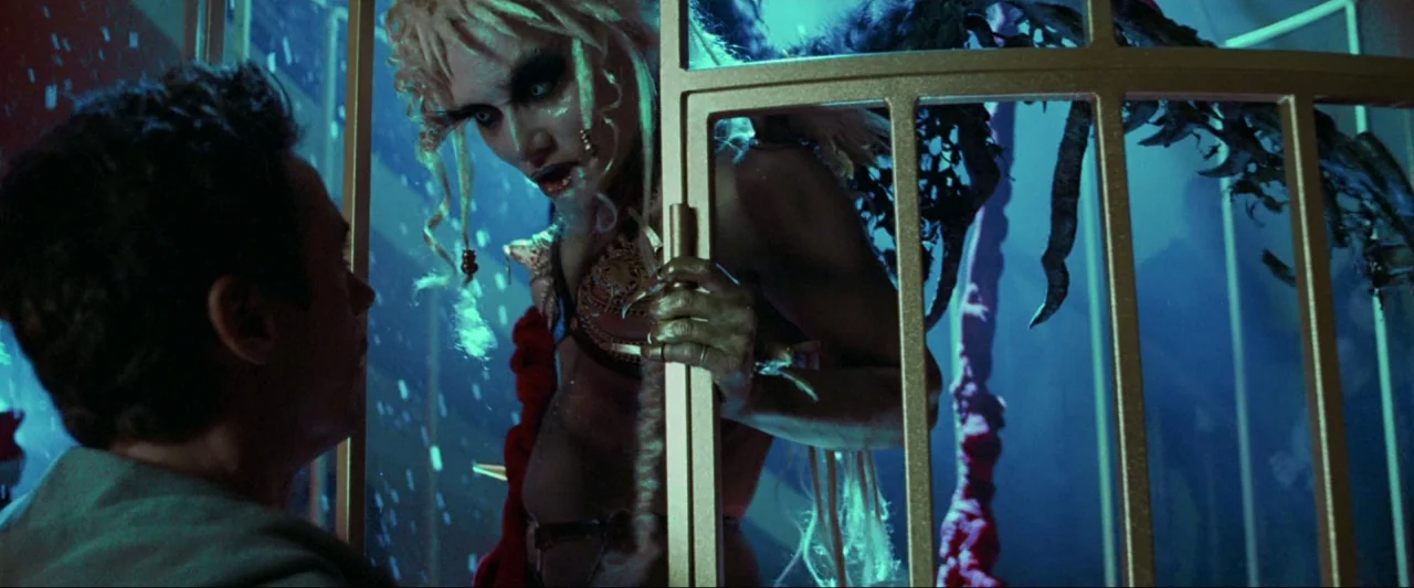 Kiss Kiss Bang Bang (2005) movie still: medium — A metal-barred cage stands within a dark, blue-lit space with scattered particles. A woman with…