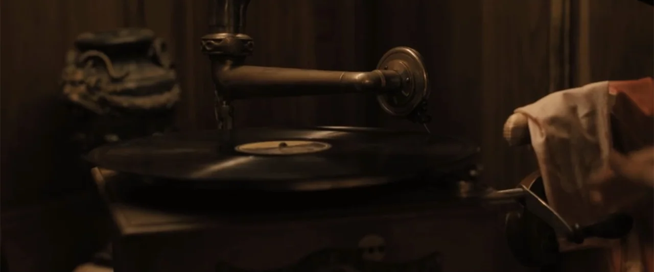 Abigail (2024) movie still: closeup — A close-up view of a vintage hand-crank phonograph sitting on a wooden surface. A black vinyl record spins on…