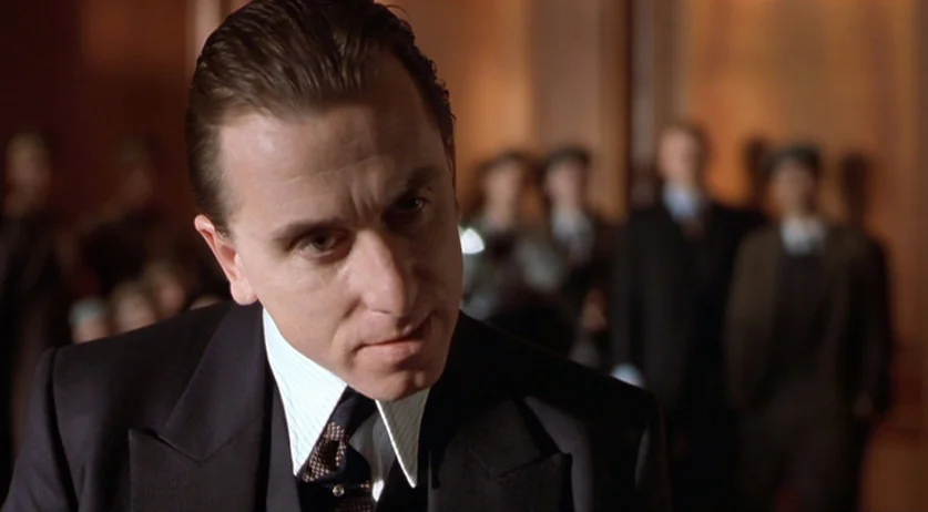 Invincible (2001) movie still: closeup — A man stands in a dimly lit, wood-paneled room among a gathering of people. He gazes slightly downward and…