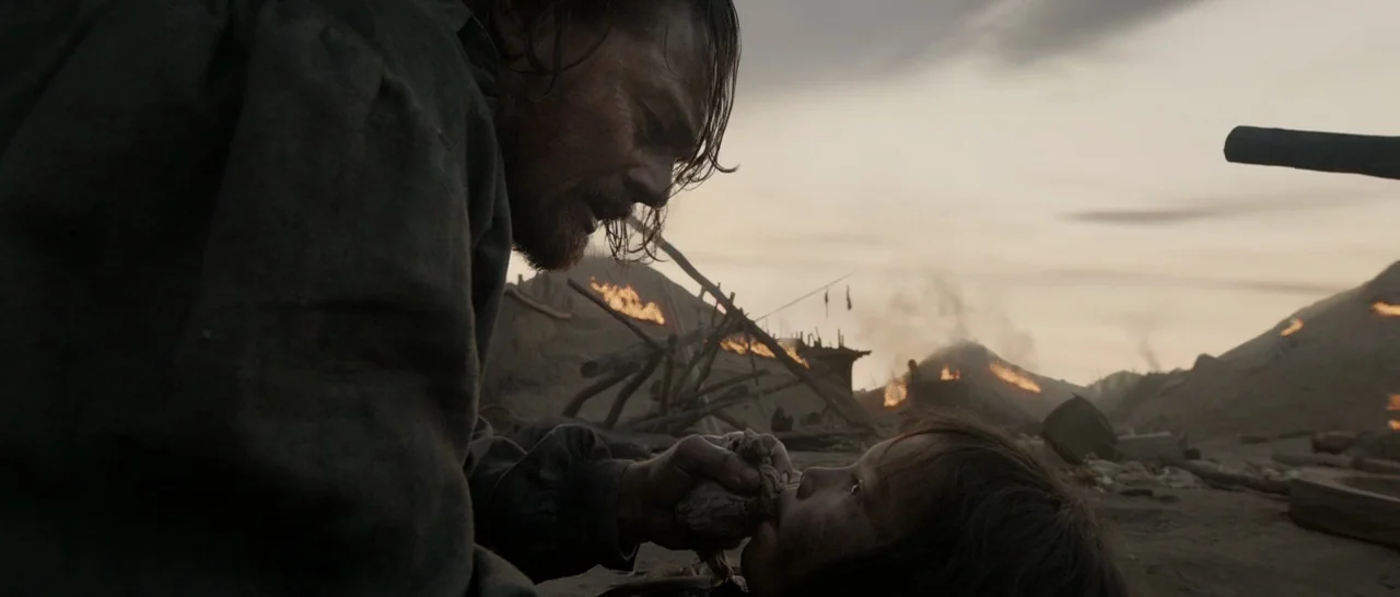 The Revenant (2015) movie still: medium — A scorched, desolate landscape features burning ruins under a dim, overcast twilight sky. A man kneels and…